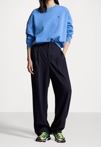 Ljust blått sweatshirt med ribbad halsringning, bärs över marinblå vida byxor. Färgglada sneakers med gröna och svarta detaljer.