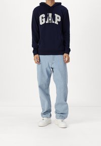 Sudadera azul marino con el logotipo "GAP" en blanco, capucha con cordón y bolsillo frontal, combinada con vaqueros holgados de un azul claro y zapatillas blancas.