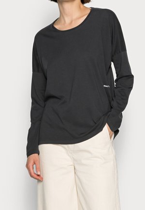 Osoba nosící volné černé tričko s dlouhým rukávem mírně zasunuté do vysokých širokých kalhot v barvě krémově bílé, s jednou rukou v kapse.