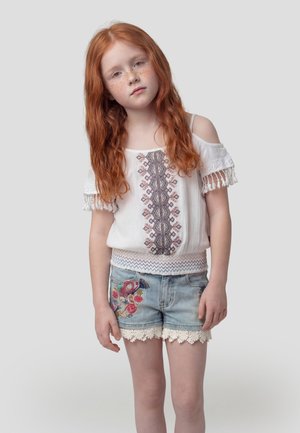 Chica pelirroja con pecas lleva una blusa blanca bordada sin hombros y shorts de mezclilla con bordado floral, de pie contra un fondo simple.