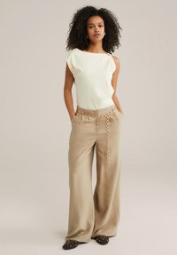 Light yellow sleeveless top with draped shoulder, paired with wide-leg beige trousers featuring a chain belt and dark slip-on shoes with studs.