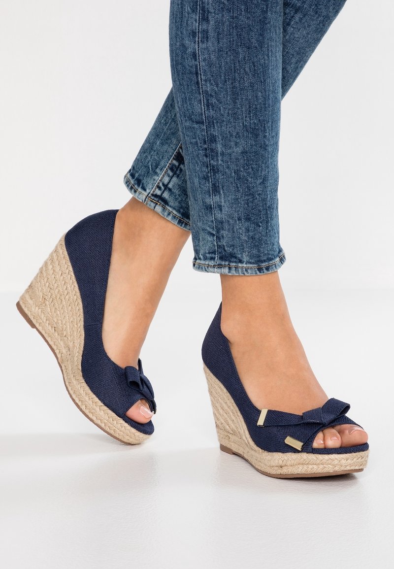 Pieds en jean denim bleu portant des sandales compensées bleu marine avec talons en corde tressée et nœuds décoratifs, posés sur une surface blanche lisse.