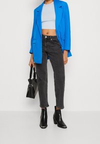 Femme portant un blazer bleu vif, un crop top bleu clair, un jean noir et des bottines noires, tenant un sac à main noir texturé sur fond blanc.