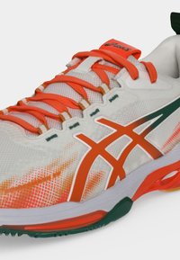 Λευκό και πορτοκαλί αθλητικό παπούτσι ASICS με πορτοκαλί κορδόνια, επάνω μέρος από δίχτυ και πράσινα και πορτοκαλί διακοσμητικά στη σόλα, εμφανίζεται από κοντινή γωνία.