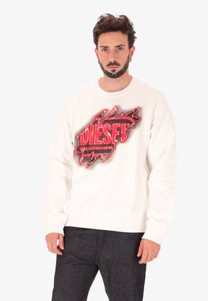 Homme portant un sweat-shirt blanc avec un texte graphique déchiré rouge et noir "DIESEL" et un pantalon sombre, se tenant devant un fond blanc uni.