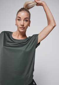 Dark green, short-sleeve athletic top with a ribbed texture and rounded neckline. Features a subtle logo tag at the hem.