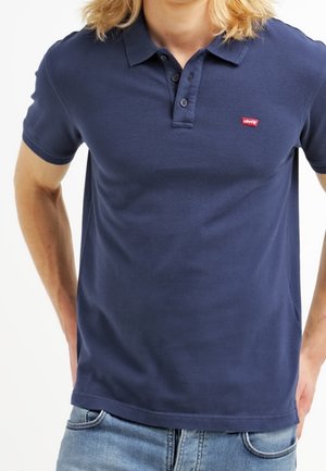 Osoba ubrana w granatową koszulkę polo Levi's z trzema guzikami i jasnoniebieskie dżinsy, ręce spoczywają na biodrach.