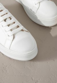 Baskets en cuir synthétique blanc avec des bouts arrondis, des lacets plats et une semelle épaisse blanche, posées sur une surface beige texturée.