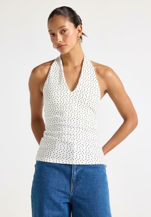 Donna che indossa un top bianco con pois neri e jeans blu a vita alta, in piedi con le braccia dietro la schiena.