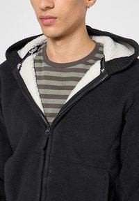 Schwarze Fleecejacke mit einem Reißverschluss vorne und einer weißen, flauschig gefütterten Kapuze. Wird über einem grau-oliv gestreiften Hemd getragen.