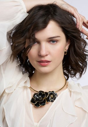 Collier de fleurs noires avec des accessoires en ton or, présentant des pétales brillants et un détail délicat. Porté sur une blouse blanche transparente.