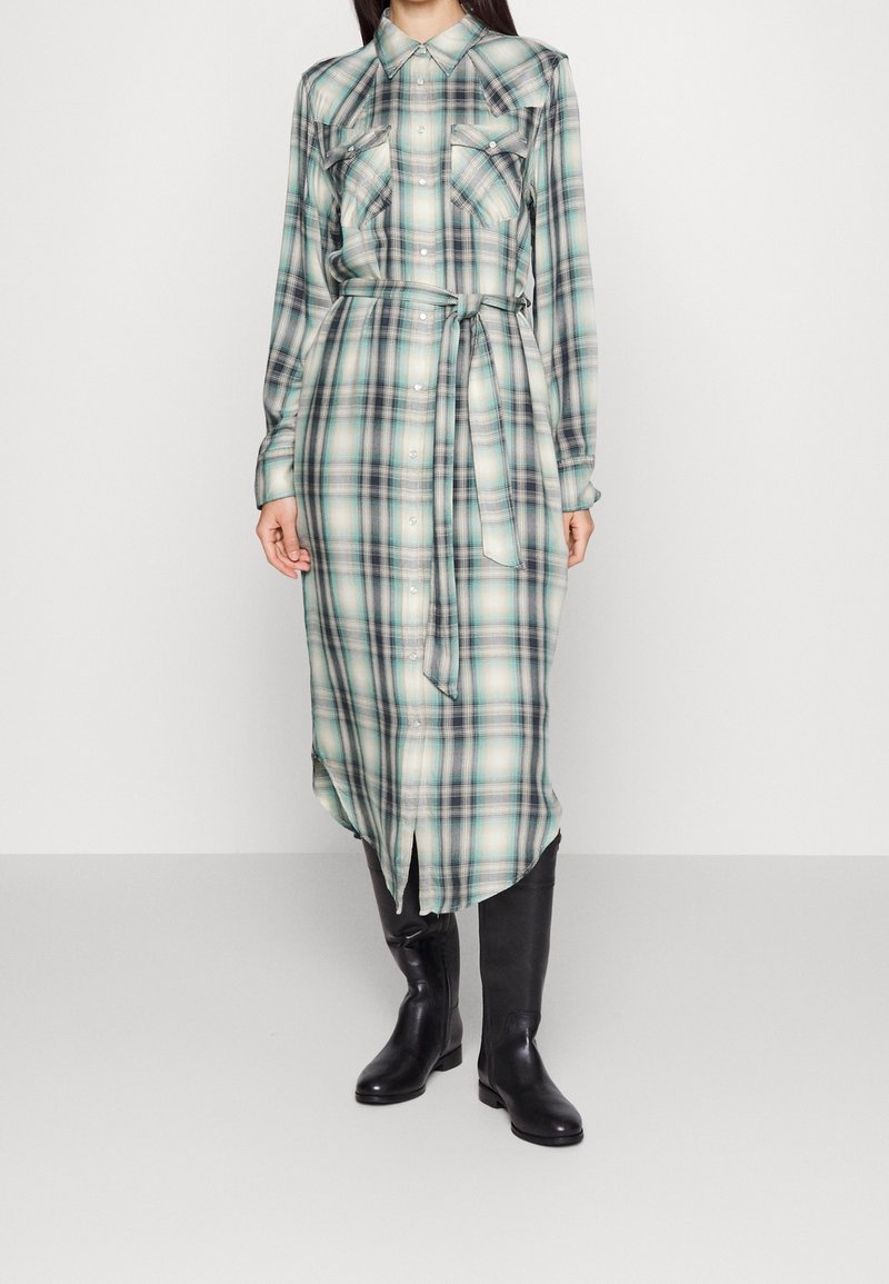 Dlouhé šaty košilového střihu s kostkovaným vzorem v zelené a modré barvě, s opaskem v pase, knoflíkovým zapínáním a kapsami. Doplněné černými kozačkami do půli stehen.