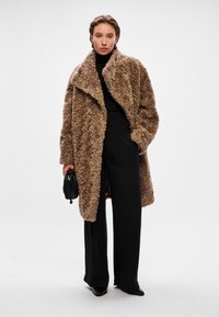 Καφέ υπερμεγέθη ψεύτικη γούνα μπουφάν με γιακά, μακριά μανίκια και υφή επιφάνειας, συνδυασμένο με μαύρα ψηλόμεσα παντελόνια.