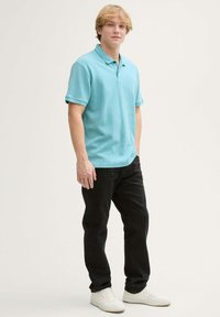 Lichtblauw polo shirt met korte mouwen, gecombineerd met zwarte jeans en witte sneakers, staand tegen een neutrale achtergrond.