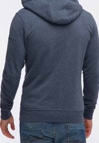 Uomo che indossa una felpa con cappuccio blu navy e jeans blu, mostrato di spalle con le mani rilassate ai fianchi.