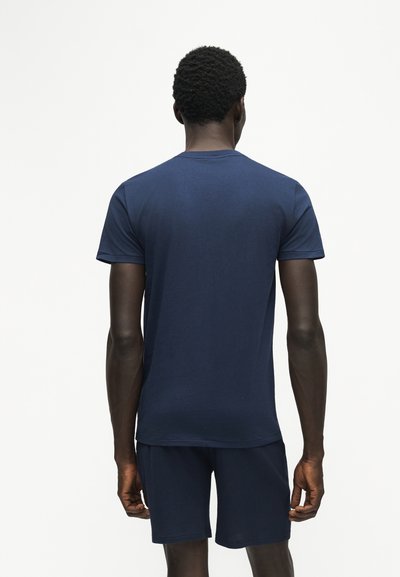 Homme debout, de dos, portant un t-shirt bleu marine uni à manches courtes et un short assorti sur fond blanc.