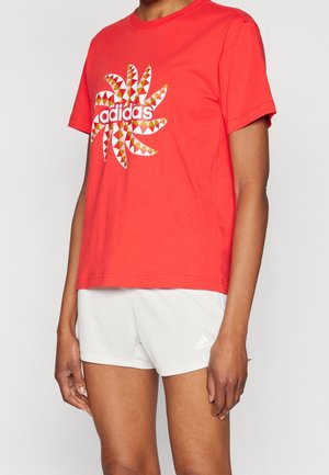 Osoba nosząca czerwoną koszulkę Adidas z kolorowym geometrycznym logo i białe sportowe spodenki Adidas.