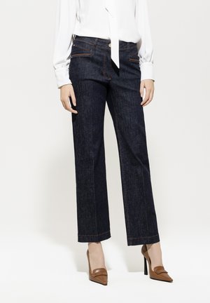 Dunkelblaue Denim-Jeans im geraden Schnitt mit orangefarbenen Nähten, vorderen Taschen und hoher Taille. Kombiniert mit einer weißen Bluse und braunen Absätzen.