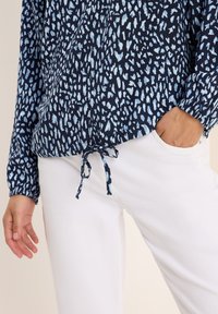 Blusa blu navy con motivo astratto azzurro, maniche arricciate e cordoncino sull'orlo, abbinata a pantaloni bianchi.