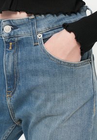 Μπλε τζιν παντελόνι ψηλής μέσης, με μπροστινή τσέπη, εμφανή ραφή και αντίθεση κίτρινη κλωστή.