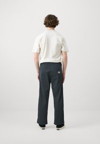 Pantalon gris foncé à coupe droite, fabriqué en tissu léger. Dispose de deux poches arrière et d'une petite étiquette de marque. Porté avec une chemise de couleur claire.