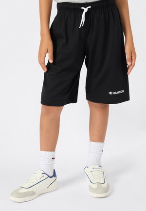 Enfant portant un short noir Champion avec un cordon blanc, des baskets blanches Champion et des chaussettes blanches, se tenant sur un fond uni.