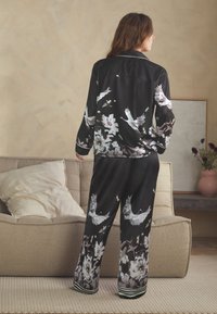 Fekete szatén pizsamaszett, fehér daru- és virágmintákkal, hosszú ujjú, gombos inggel és csíkos részletekkel díszített bő szabású nadrággal.