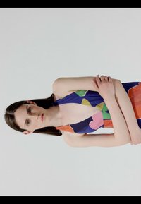 Jeune femme aux longs cheveux bruns, debout devant un fond uni, portant une robe sans manches aux motifs colorés avec un profond décolleté en V.