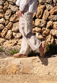 Sandales plates en cuir beige avec une large bande, semelle en caoutchouc et un léger talon. Portées avec un pantalon en lin ample devant un mur en pierre.