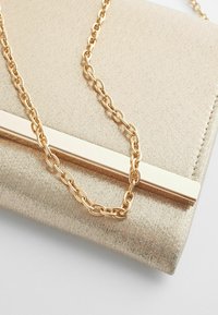 Pochette métallisée dorée avec un détail lisse en barre métallique dorée et une grosse chaîne dorée reposant sur la pochette.
