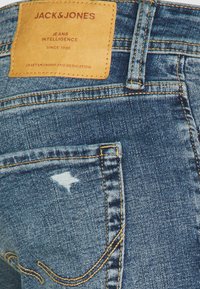 Bakh pocket på blå denimjeans med en brun läderlogga som säger "JACK & JONES" och en liten sliten textildetalj på fickan.