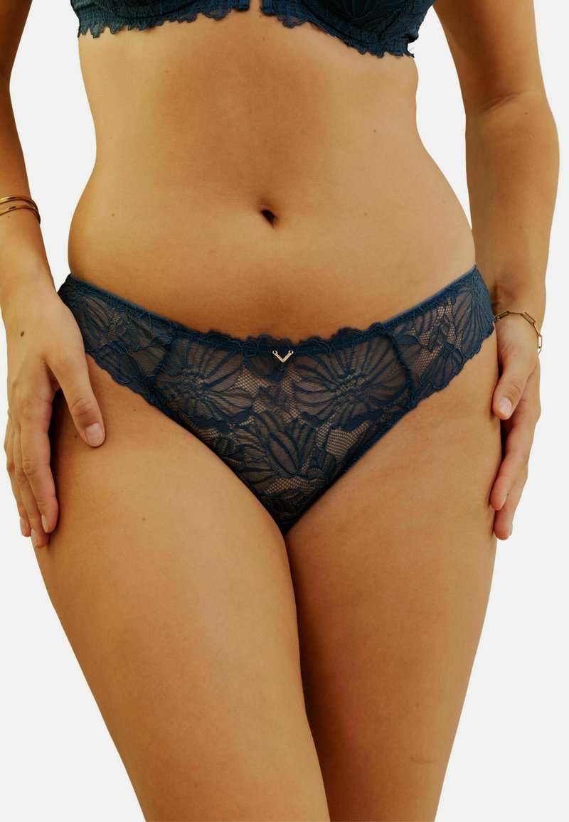 Culotte hipster en dentelle de couleur bleu sarcelle avec des motifs floraux et un bord festonné. Présente un petit détail en forme de cœur métallique au centre devant.