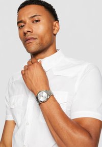 Sudraba rokas pulkstenis ar apaļu seju un romiešu cipariem, apvienots ar baltu īso piedurkņu krekliņu ar krūšu kabatām.