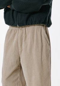 Beige kordbársony nadrág bordázott textúrával, elasztikus derékrésszel és oldalsó zsebekkel, fekete, testhezálló kabáttal párosítva.