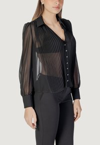 Blusa nera trasparente con plissé verticali, apertura frontale con bottoni e colletto classico. Presenta maniche lunghe e morbide con polsini aderenti.