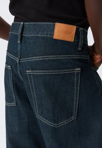 Tamnoplave traperice od trapera sa smeđom kožnom oznakom na kojoj piše "WEEKDAY." Imaju klasične džepove i detalje šavova duž šavova.