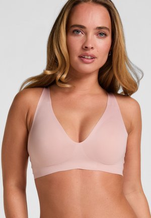 Frau mit langen blonden Haaren, die einen hellrosa nahtlosen BH trägt, steht vor einem schlichten grauen Hintergrund und schaut geradeaus.