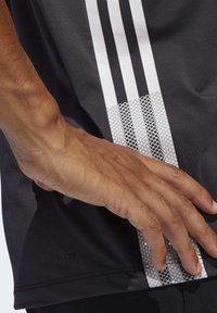 Černé sportovní tričko s třemi bílými svislými pruhy a texturovanou síťovinou s kulatými výřezy blízko lemu.