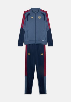 Survêtement de sport bleu et marine avec des bandes rouges, veste zippée intégrale et pantalon assorti arborant les logos dorés d’Adidas et du Bayern Munich.