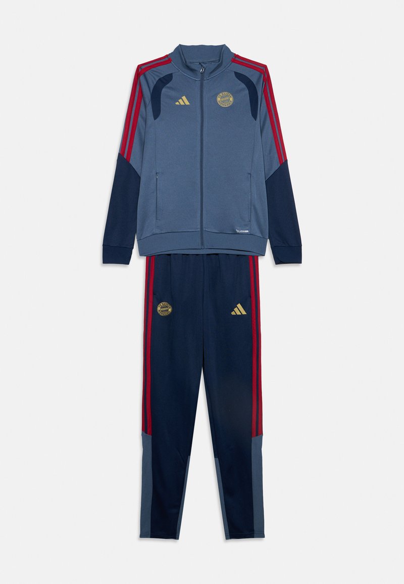 Survêtement de sport bleu et marine avec des bandes rouges, veste zippée intégrale et pantalon assorti arborant les logos dorés d’Adidas et du Bayern Munich.