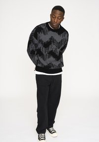 Schwarzer und grauer gemusterter Strickpullover mit Rundhalsausschnitt, über einem weißen Hemd getragen. Kombiniert mit lockeren schwarzen Hosen und schwarzen Sneakers.