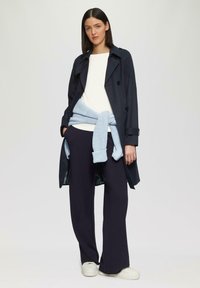 Manteau trench navy, haut blanc, pantalon large navy, et pull bleu clair noué à la taille ; des chaussures blanches complètent la tenue.