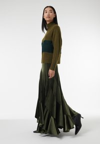 Groene gebreide trui met een teal-streep, gecombineerd met een luchtige, donkergroene satijnen maxi-rok met gelaagde ruffles en zwarte enkellaarsjes.