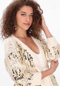 Kremowy kardigan z złotymi i srebrnymi cekinami, otwarty z przodu, z prążkowanymi mankietami i paskiem w talii. Miękka tekstura i luźny krój.