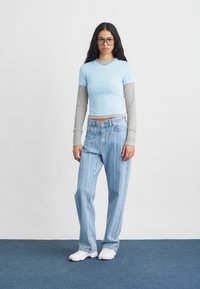 Γυναίκα με μακριά μαύρα μαλλιά και γυαλιά φοράει στρωματοποιημένα μπλε και γκρι μπλουζάκια, ανοιχτό μπλε ριγωτά τζιν και λευκά αθλητικά παπούτσια, ενώ στέκεται σε μπλε χαλί.