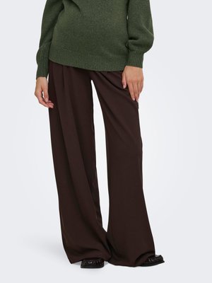 Personne portant un pantalon large marron foncé, des chaussures noires et un pull en laine vert, debout devant un fond uni.