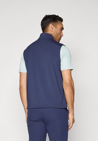 Man facing away wearing navy sleeveless vest over light blue short-sleeve shirt and matching navy pants against plain background.