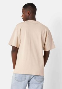 Camiseta de algodón beige con un corte relajado, mangas cortas, escote redondeado y un sutil detalle de logo en la parte superior de la espalda. Combinada con jeans de color azul claro.