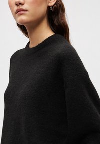 Μαύρο πλεκτό πουλόβερ με στρογγυλή λαιμόκοψη και μακριά μανίκια. Το ύφασμα φαίνεται απαλό και υφασμένο, με άνετη εφαρμογή.