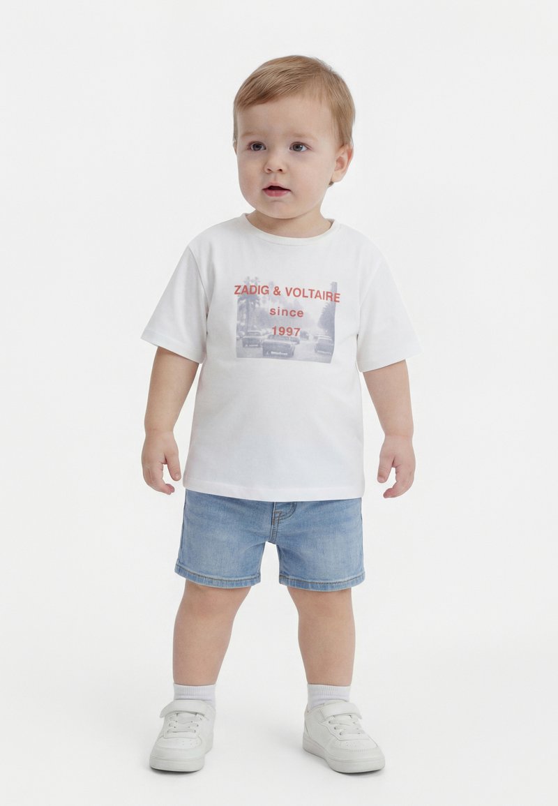 Νήπιο με ξανθά μαλλιά που φοράει λευκό μπλουζάκι με σχέδιο, ανοιχτόχρωμα τζιν σορτς, λευκές κάλτσες και λευκά αθλητικά παπούτσια, στέκεται μπροστά σε απλό φόντο.